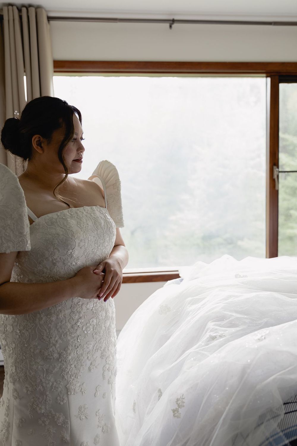 Bride in off-shoulder white lace wedding gown stands pensively by a window with soft natural light.