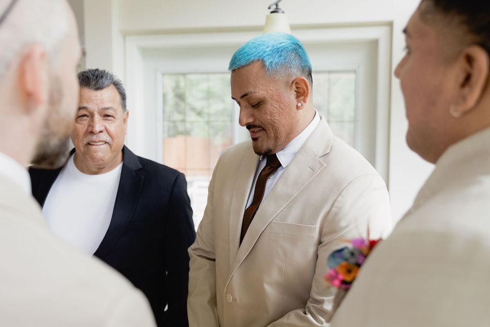 Group of men in suits converse indoors, one with distinctive blue-dyed hair at a wedding celebration.