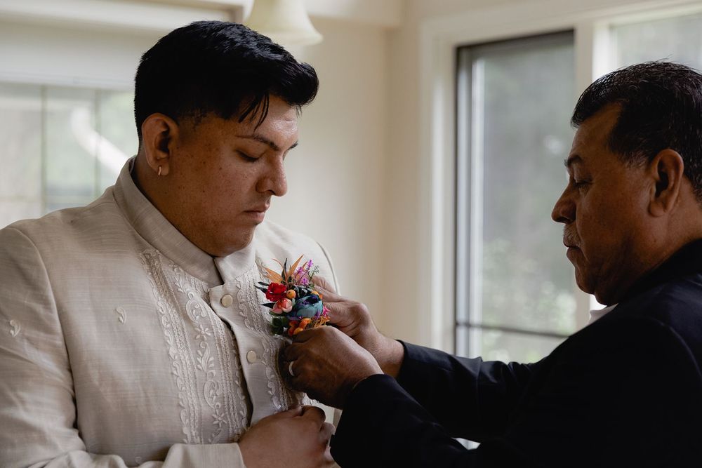 An older man pins a colorful boutonniere onto a groom wearing a cream barong tagalog before a wedding ceremony.