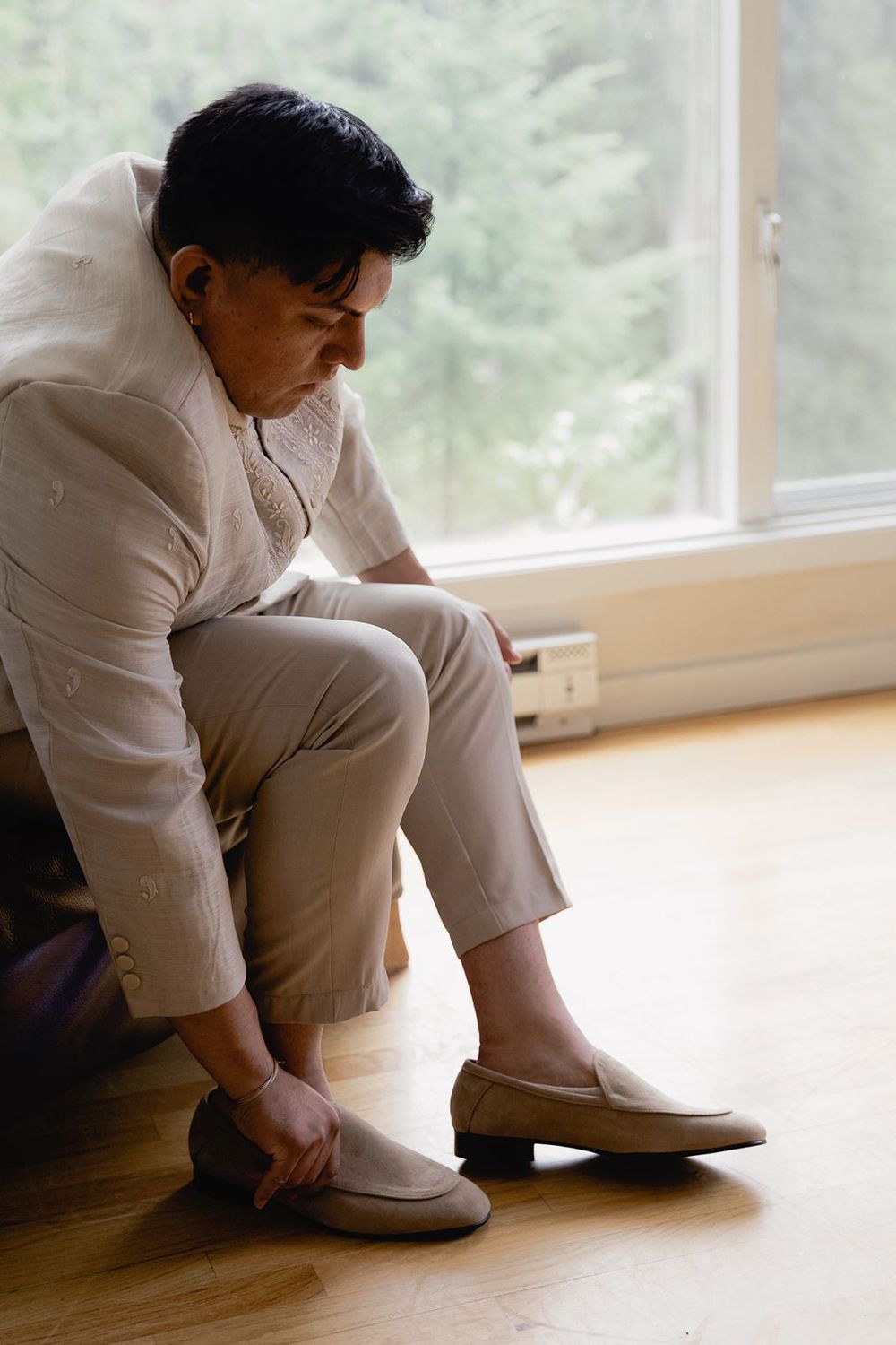 A groom in a cream barong tagalog sits on the floor putting on beige loafers before his wedding ceremony.