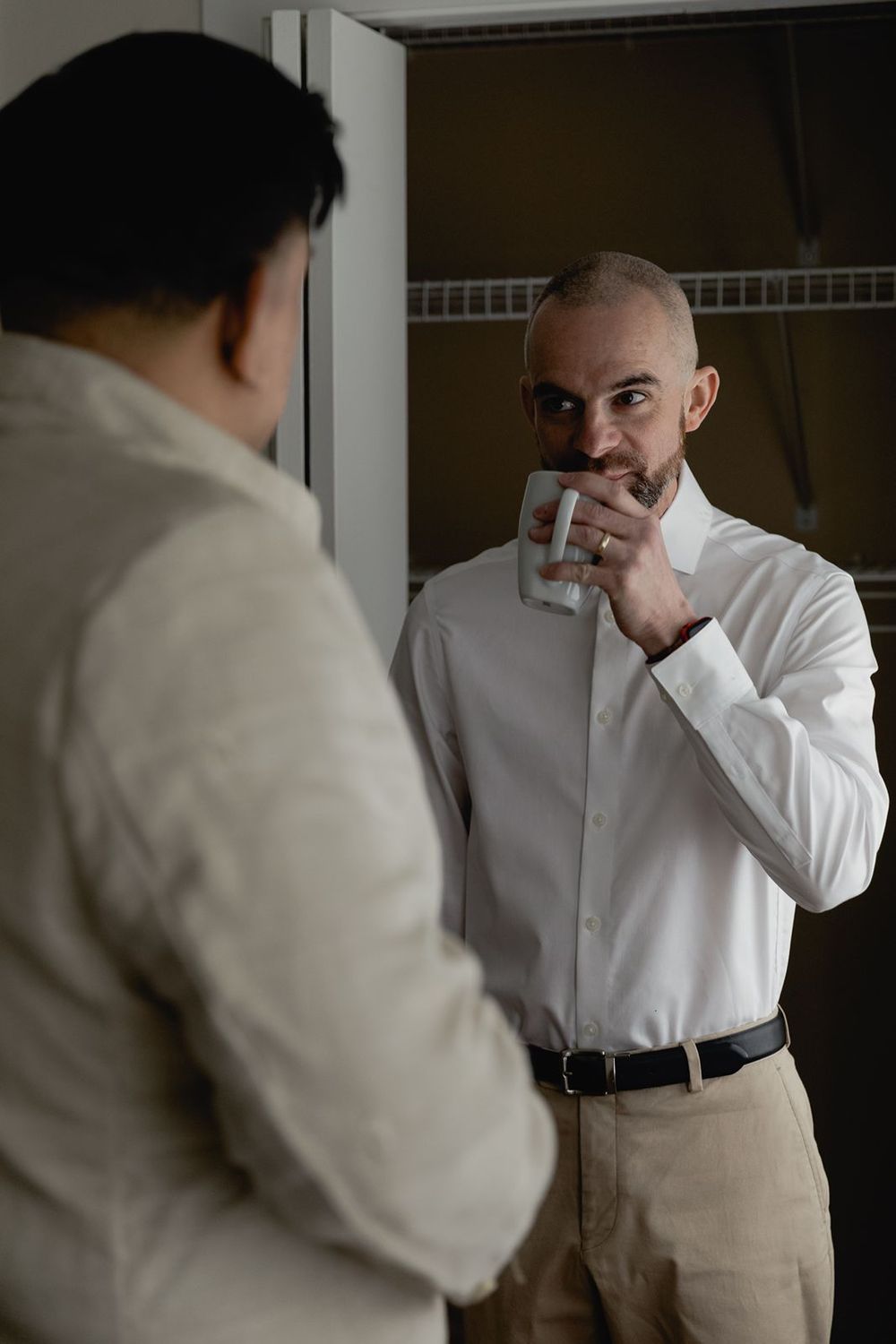 A bald groomsman in a white dress shirt drinks from a mug while chatting with a groom before the wedding.