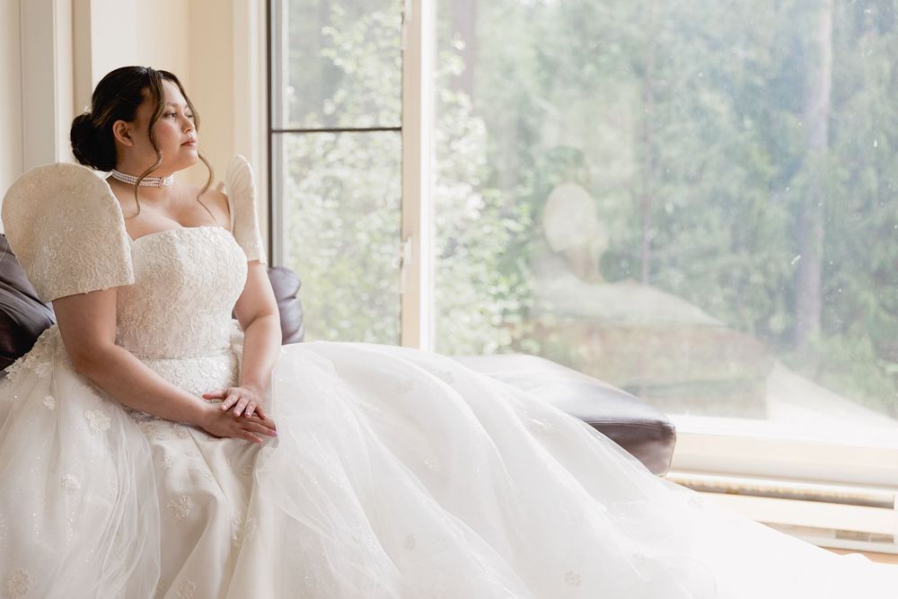 Bride in a white ball gown sits by a large window, gazing outside with a serene expression.