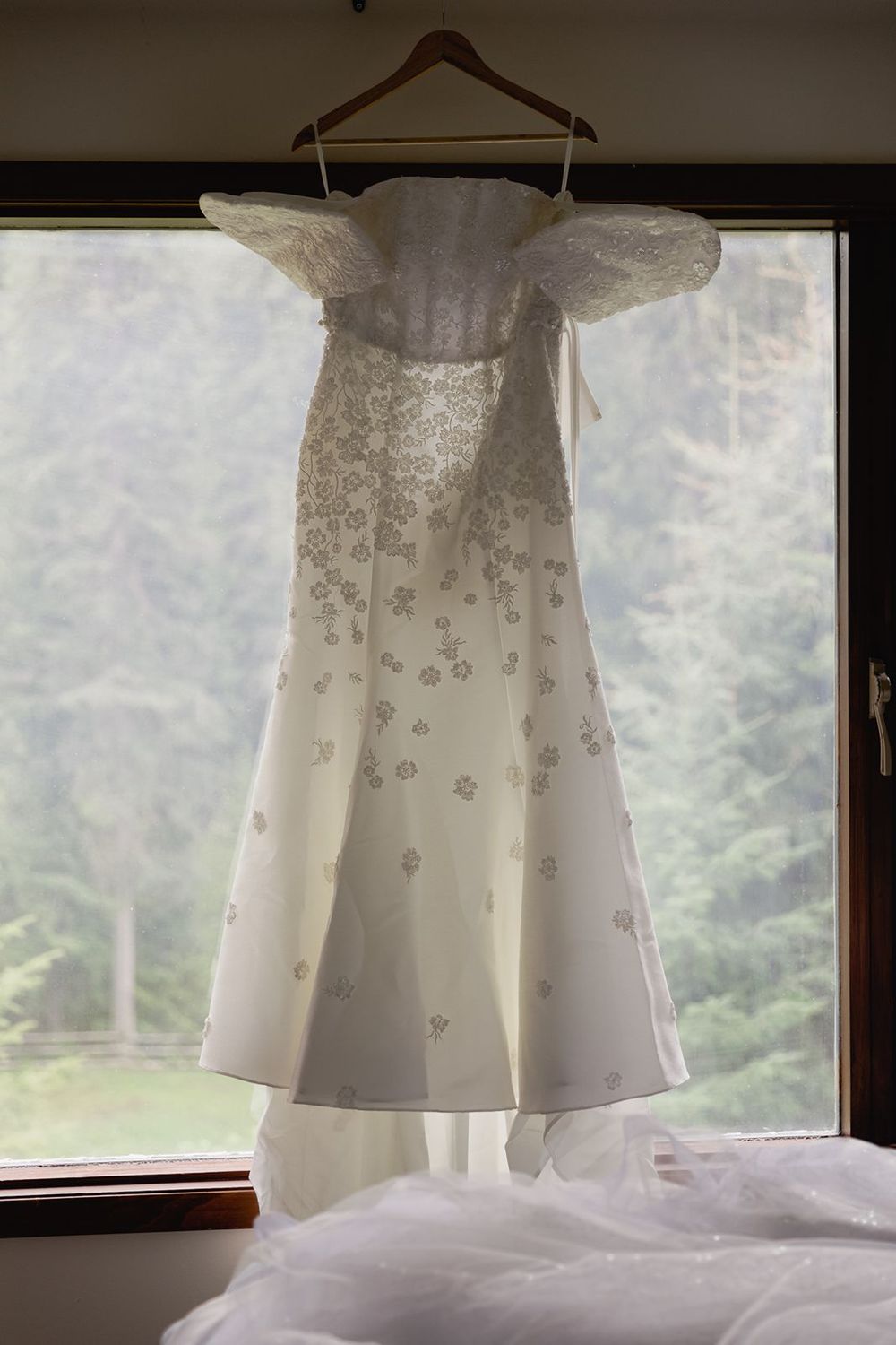 Elegant white off-shoulder wedding dress with floral embroidery hanging by a window with forest view.