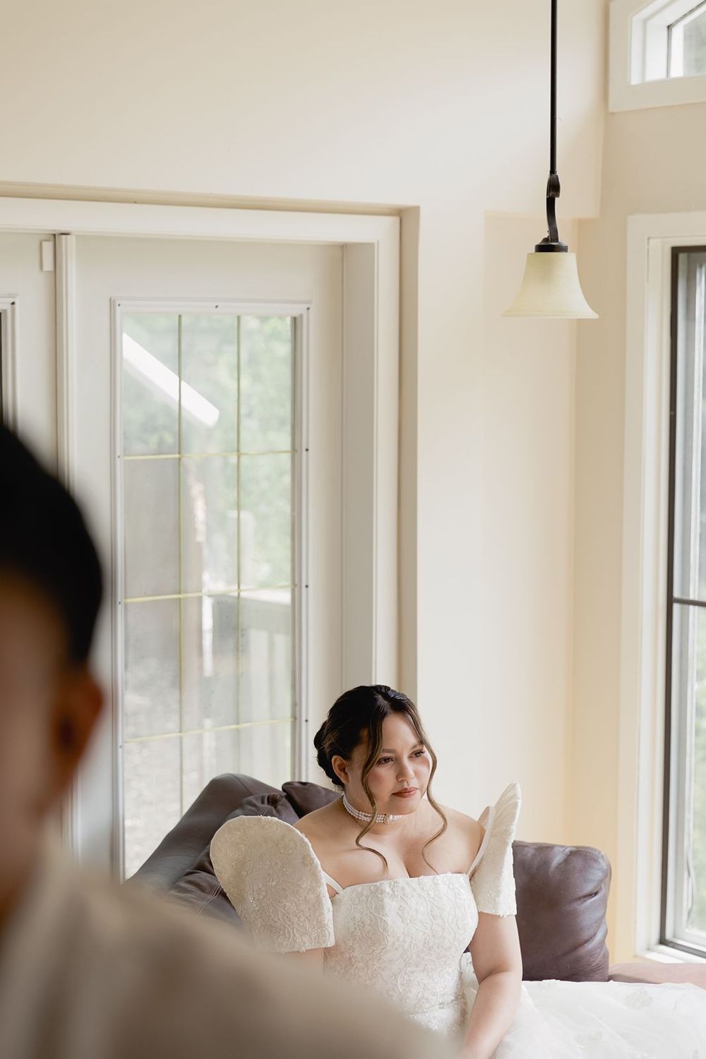 Bride in white strapless lace gown sitting on a sofa in a bright room before her wedding ceremony.