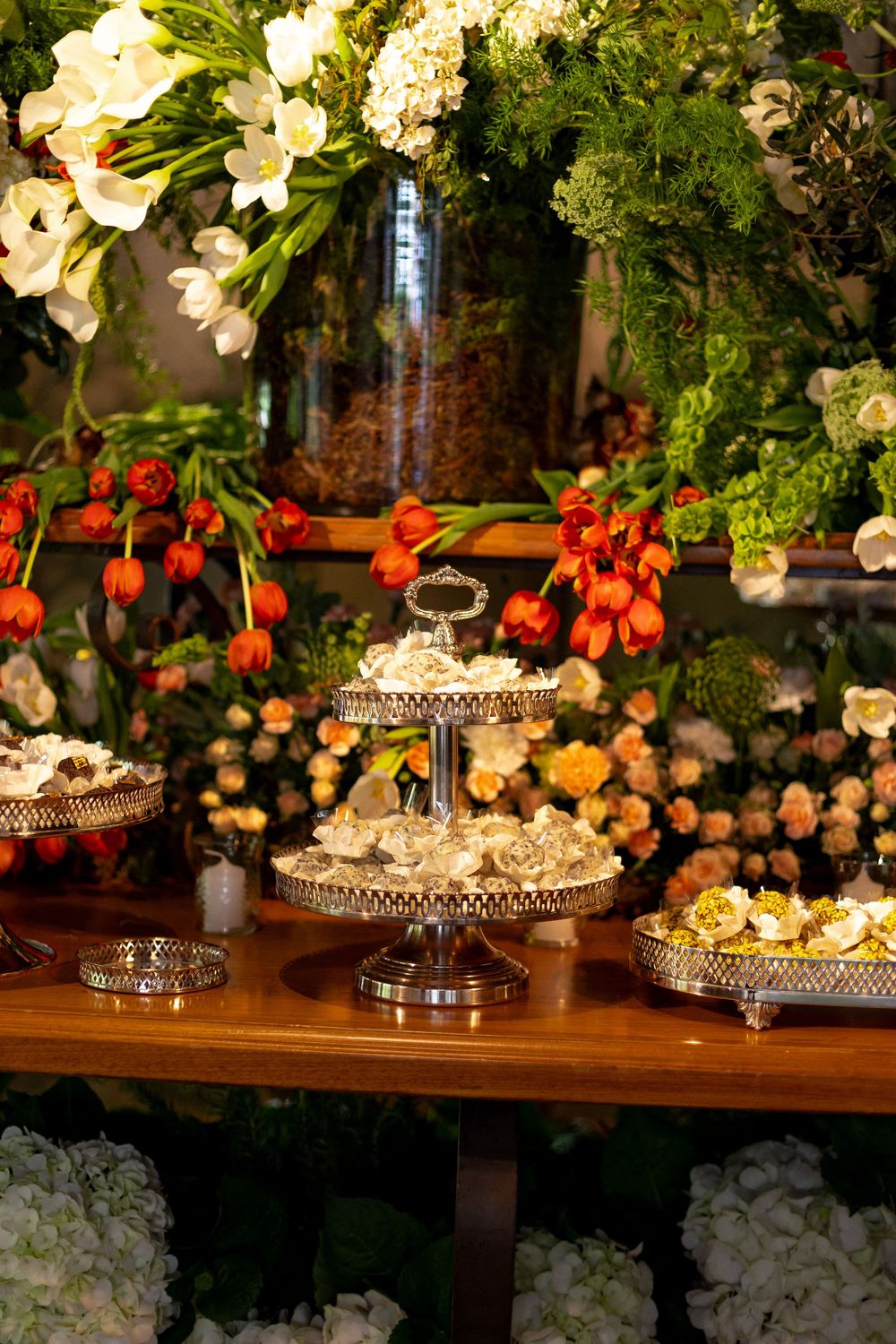 Ornate silver cake stands and floral arrangements with white blooms and red accents decorate a wooden table setting.