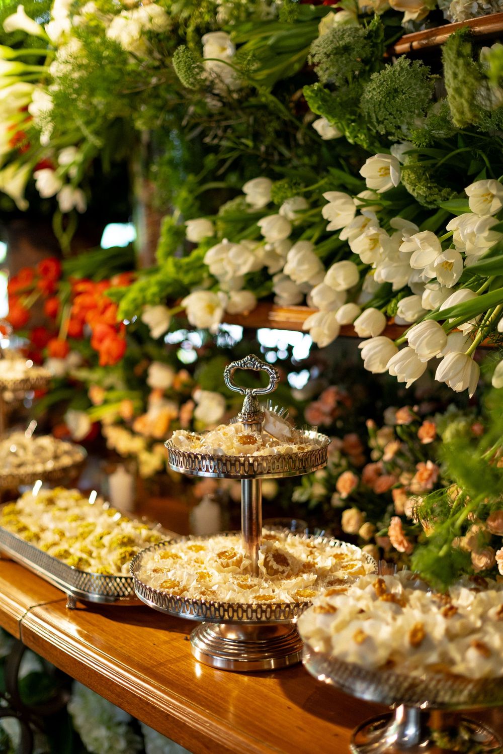 Elegant dessert display with white flowers and ornate silver serving platters on a wooden table.