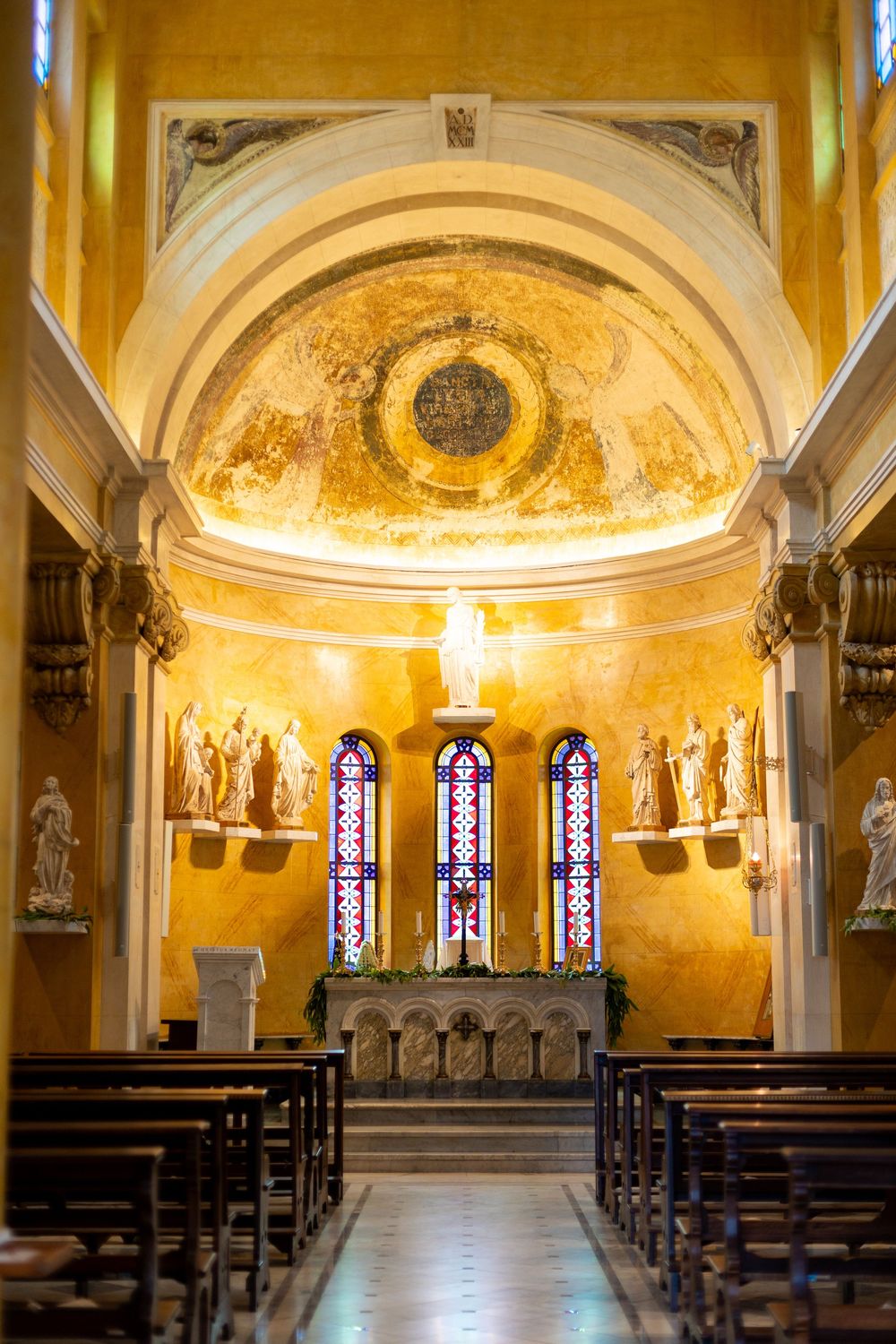 Interior of a Catholic church with golden vaulted ceiling, stained glass windows, and white statues along the walls.
