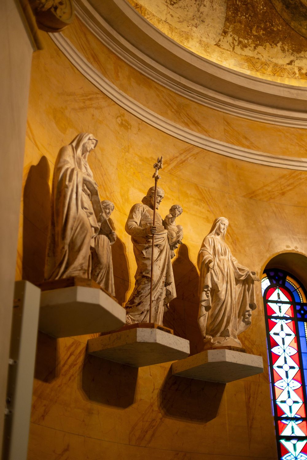 Religious statues and stained glass windows line the curved wall of a church with warm golden lighting.
