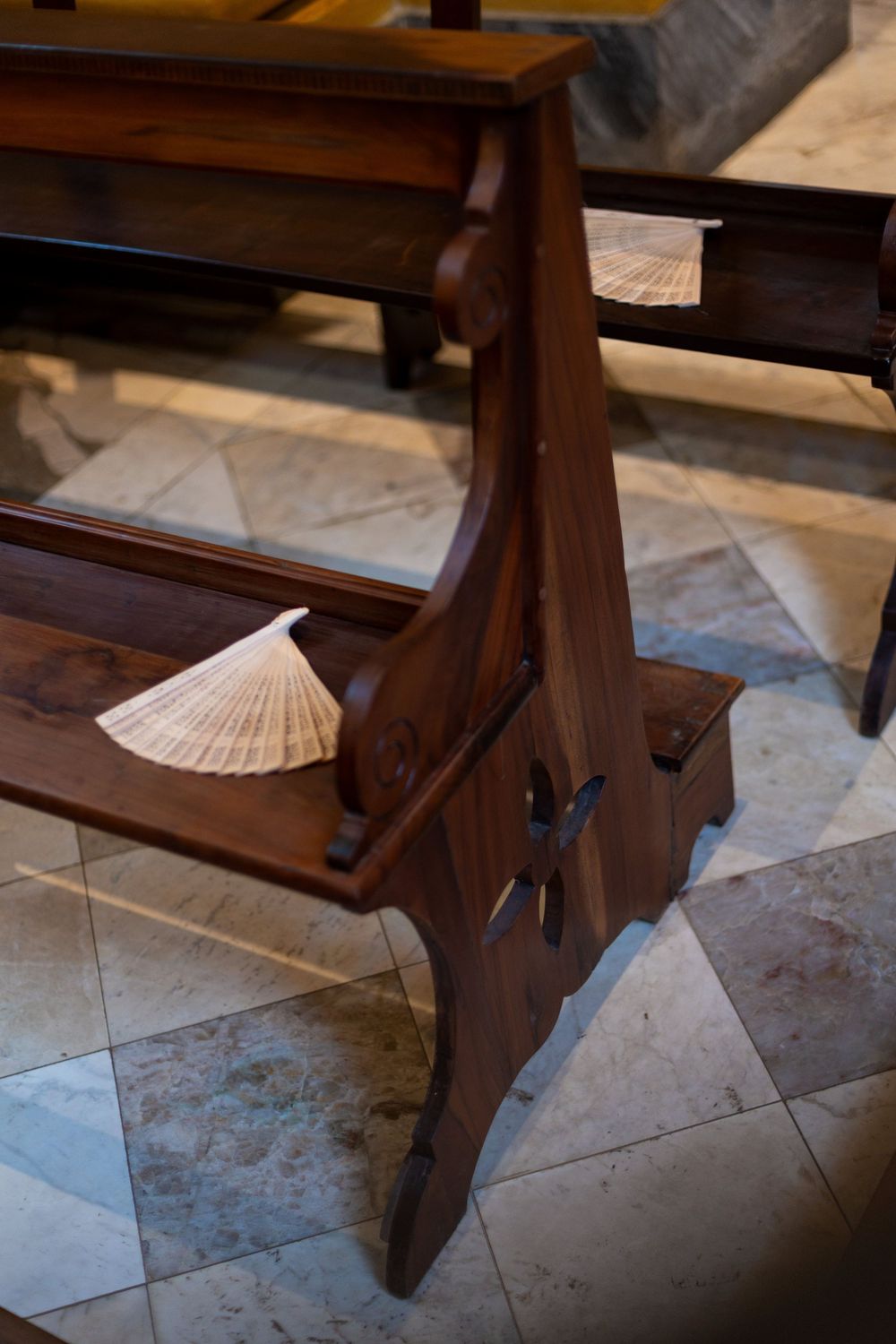 A wooden church pew with decorative details and a paper fan resting on its surface in a marble-floored space.