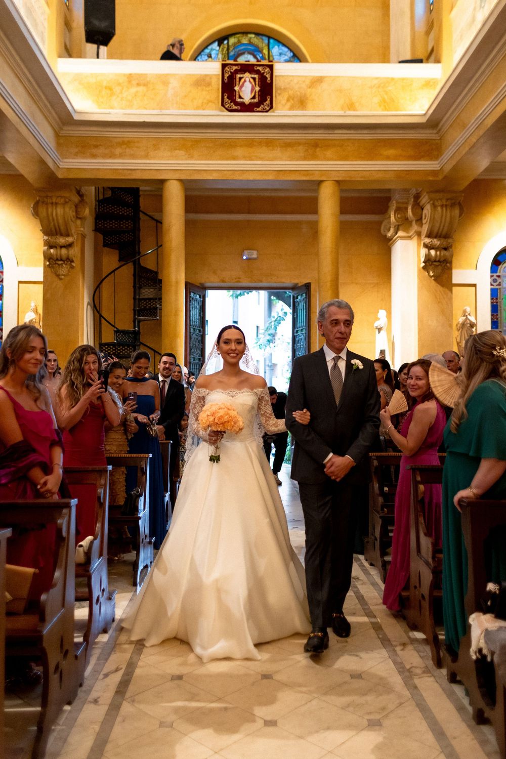 Wedding guests line the aisle of a grand yellow-walled venue as the bride processes down in her white gown.