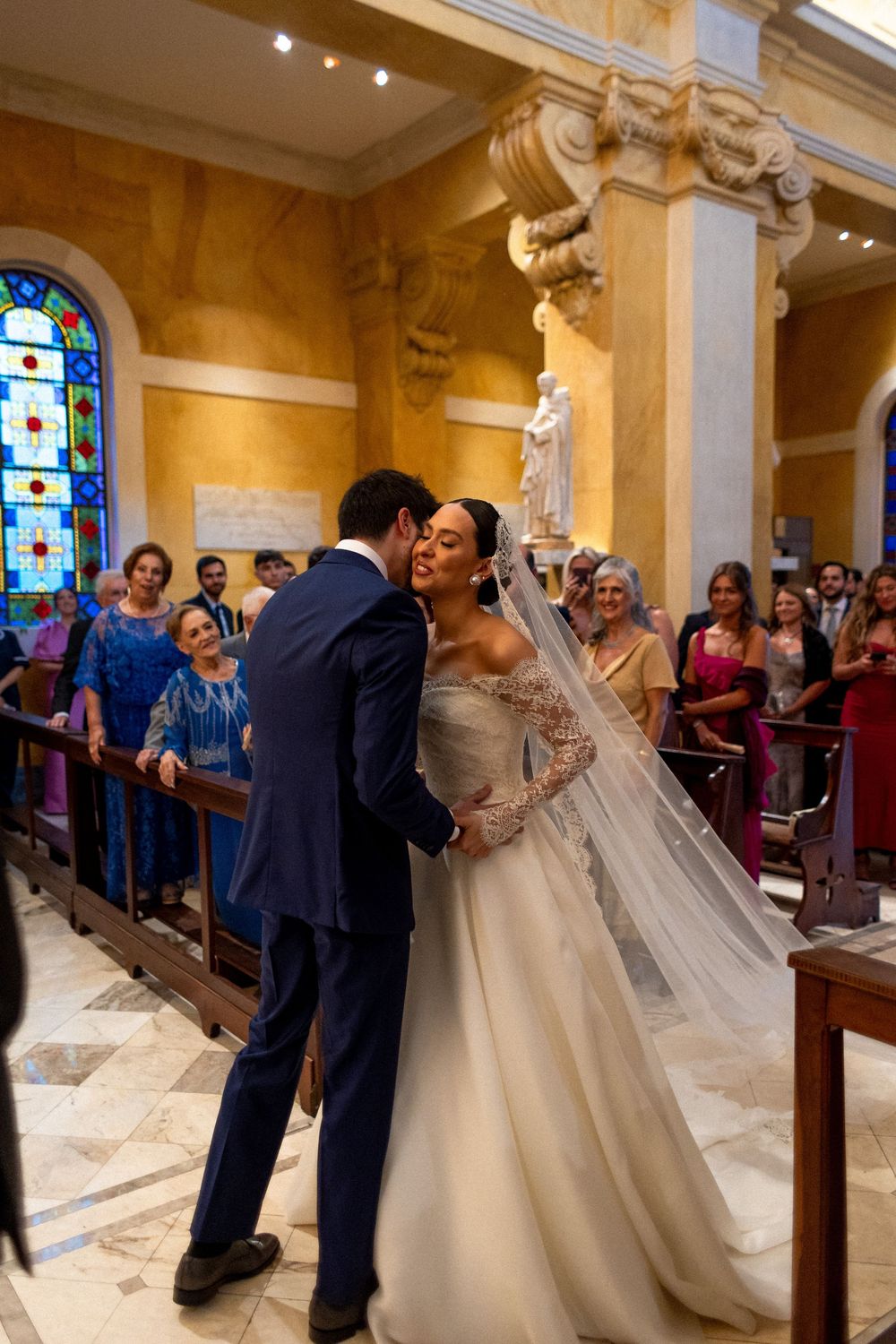 A bride in a flowing white gown dances with guests in a grand church hall with stained glass windows.