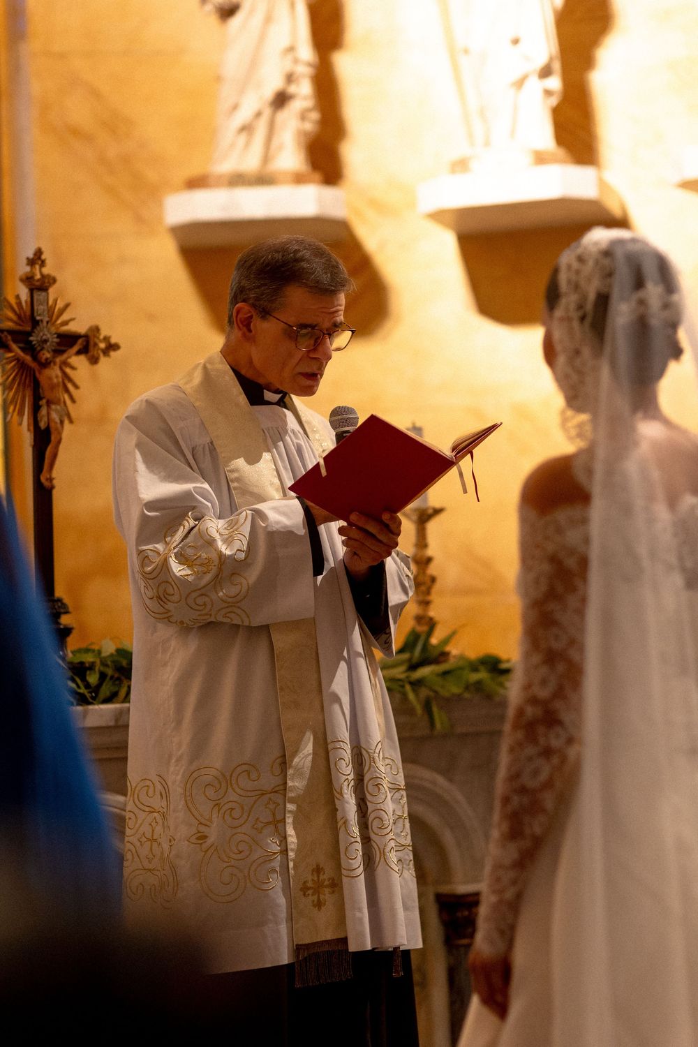A priest in white robes conducts a wedding ceremony in a warmly lit church interior.