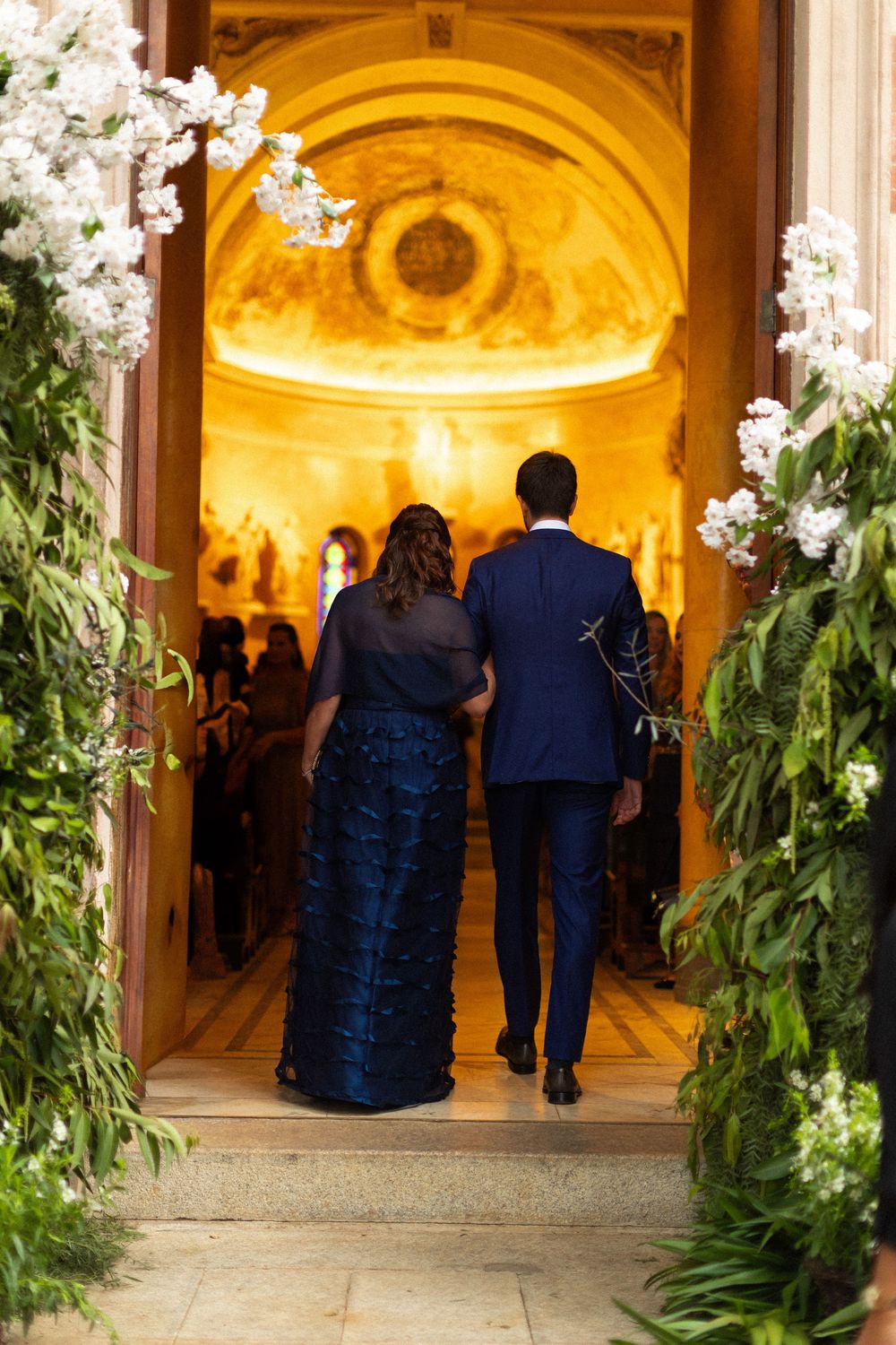 A couple walks through a floral-lined church entrance with golden lighting and white blossoms framing the doorway.