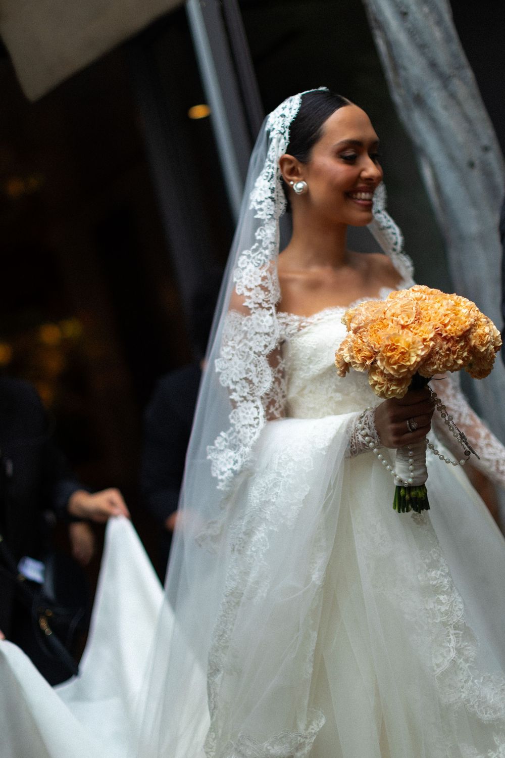 A bride holds a vibrant orange bouquet while wearing an intricate lace veil and white wedding gown.