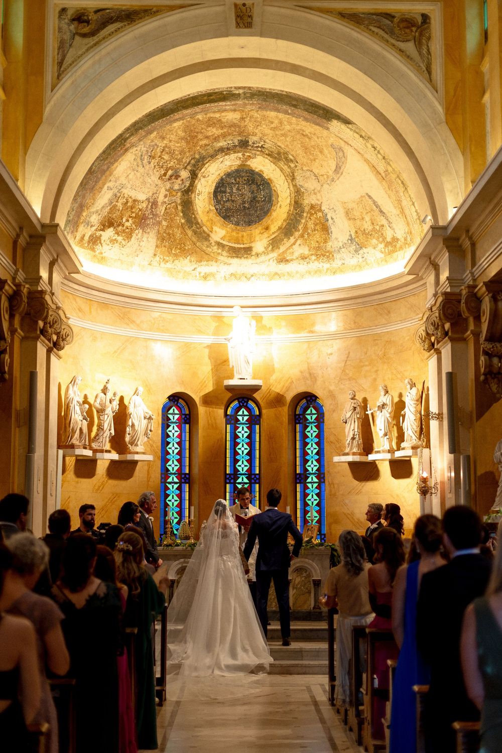 Wedding ceremony in an ornate cathedral with stained glass windows and vaulted ceilings illuminated in warm light.