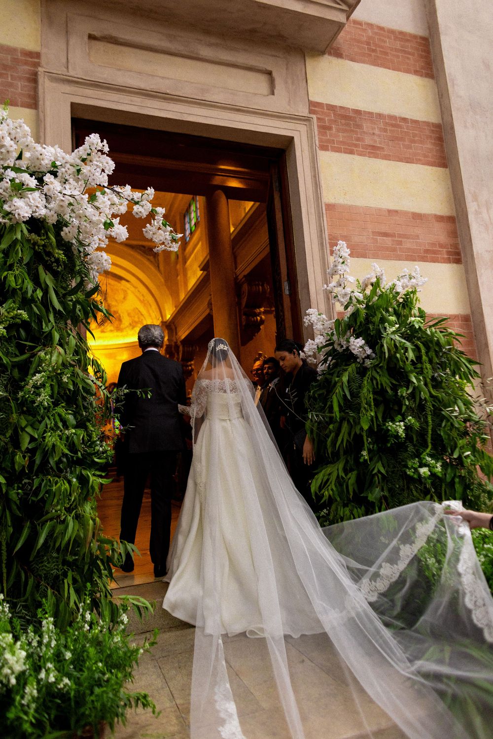 A bride in white gown enters church doors flanked by lush green plants and floral arrangements.