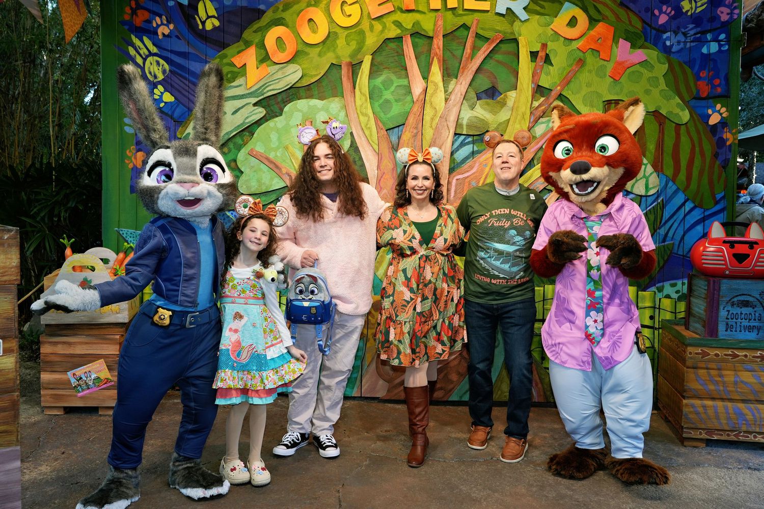 A group poses with costumed characters at Zootopia Land in a colorful theme park setting with magical forest backdrop. Zootopia