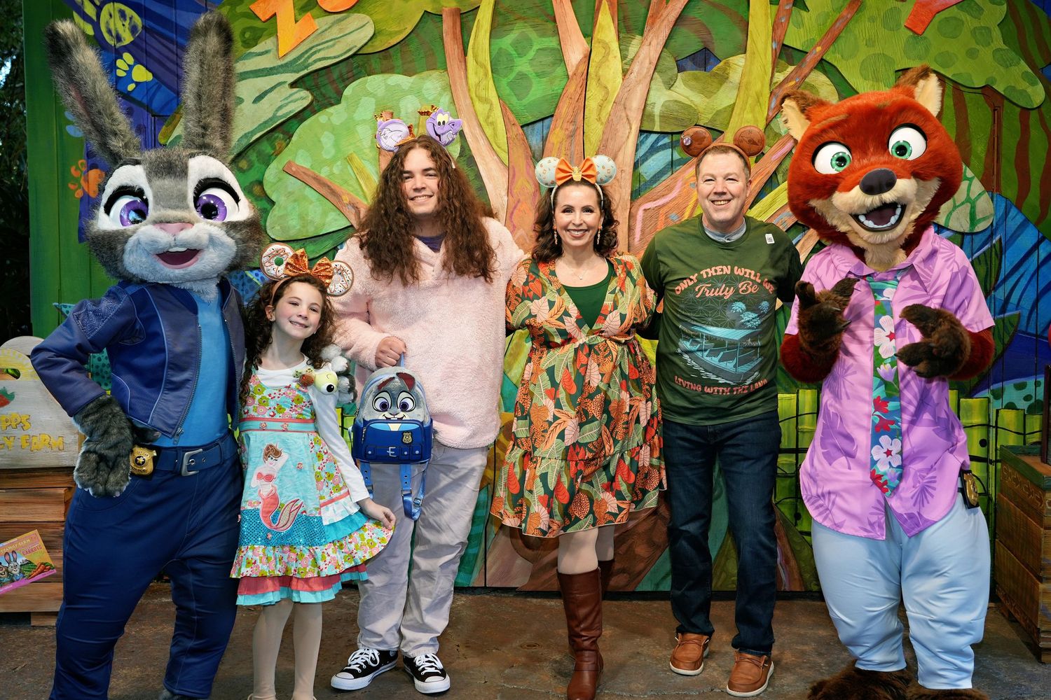 A group poses with colorful cartoon animal mascot characters against a vibrant painted forest backdrop.
