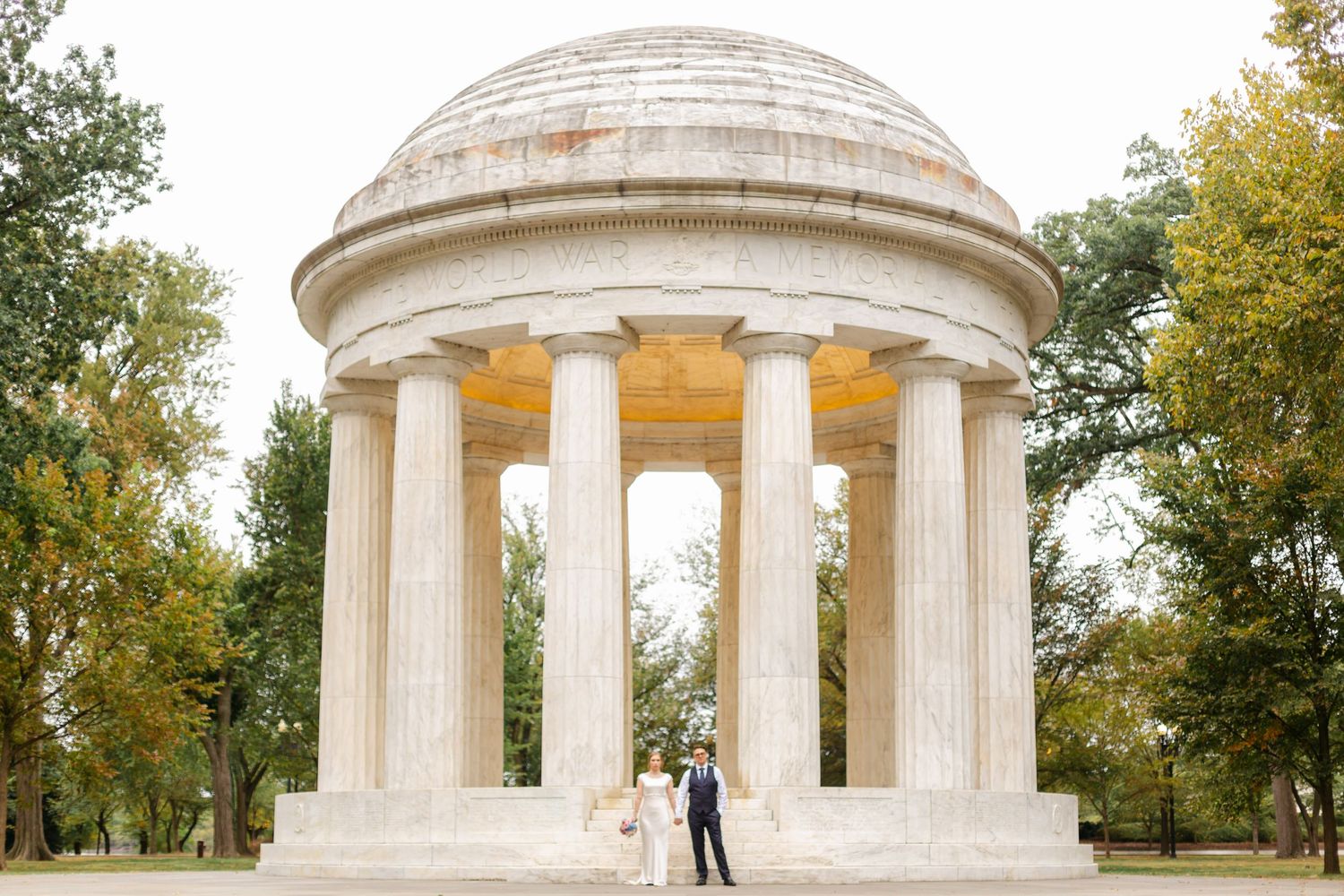 Wedding couple standing beneath grand white domed rotunda with classical columns in scenic park setting.