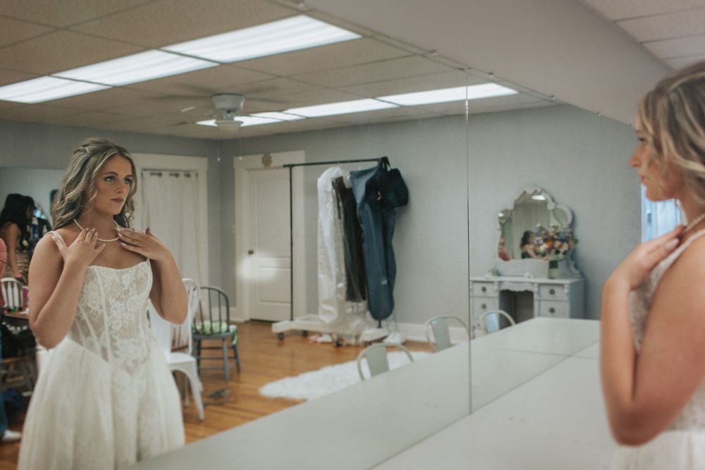 A bride in a white lace gown looks at her reflection in a dressing room mirror.