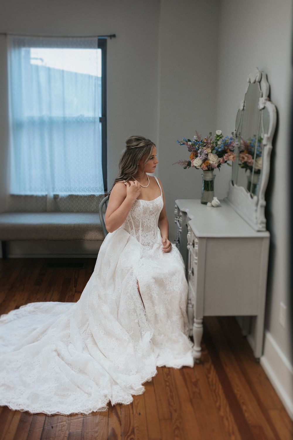 A bride in a flowing white lace gown stands beside a window and dresser with flowers.