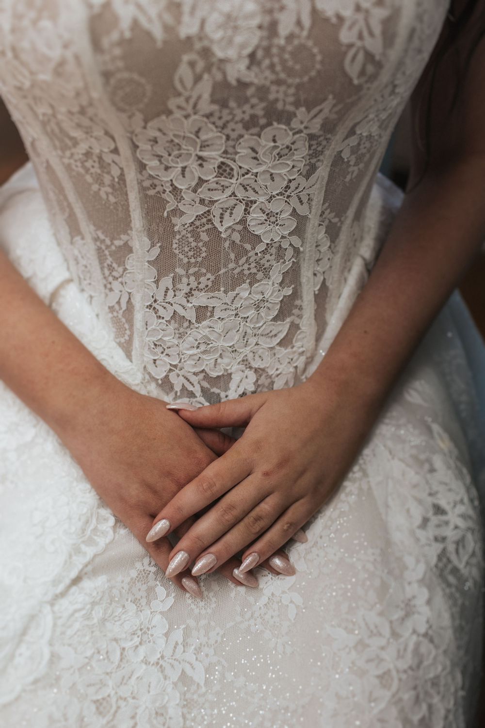 Close-up detail of intricate lace pattern and folded hands on a white wedding dress.