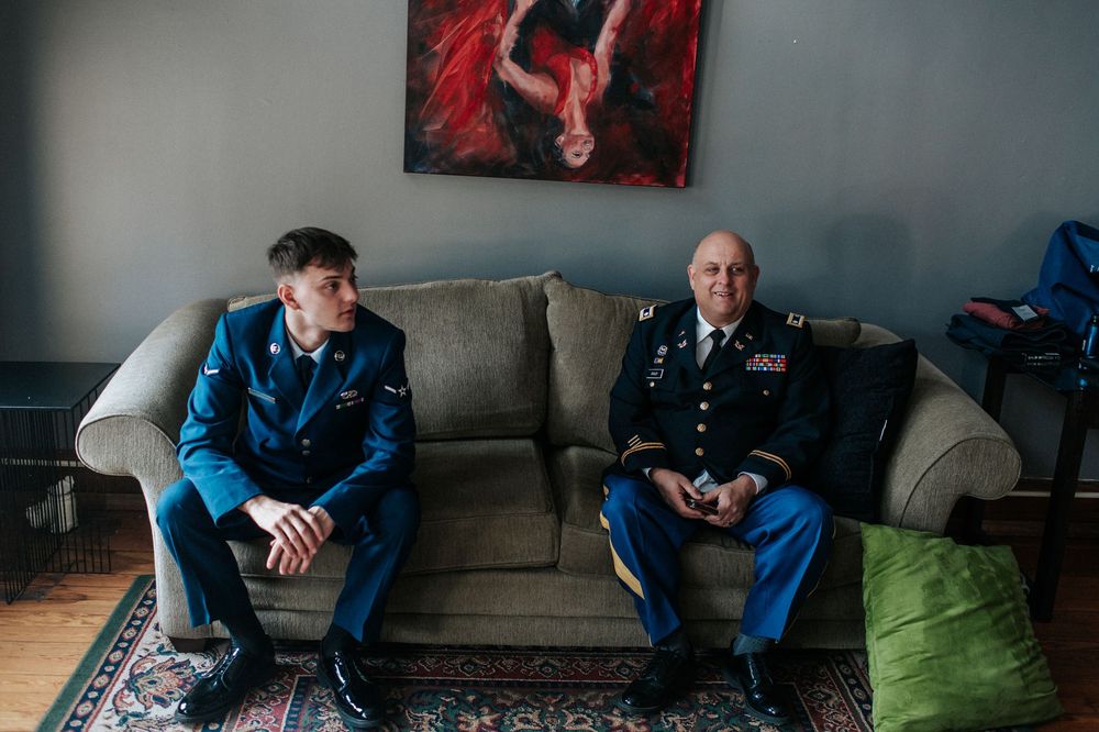 Two military service members sit on a gray couch beneath red artwork.