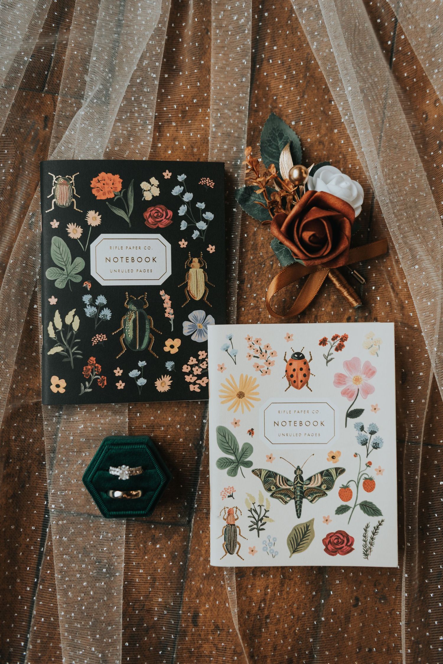 Decorative notebooks with botanical illustrations displayed alongside emerald green ring boxes on dark wood.
