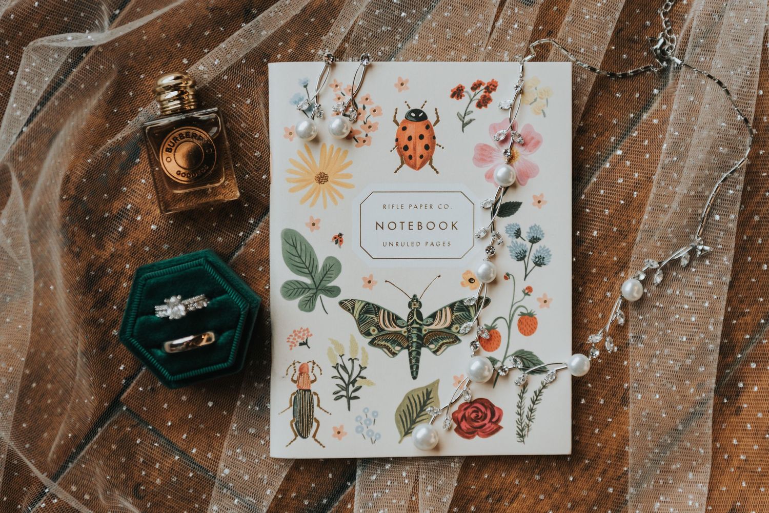 A floral and insect themed notebook lies on a wooden surface with a vintage lantern and green glass ornament.