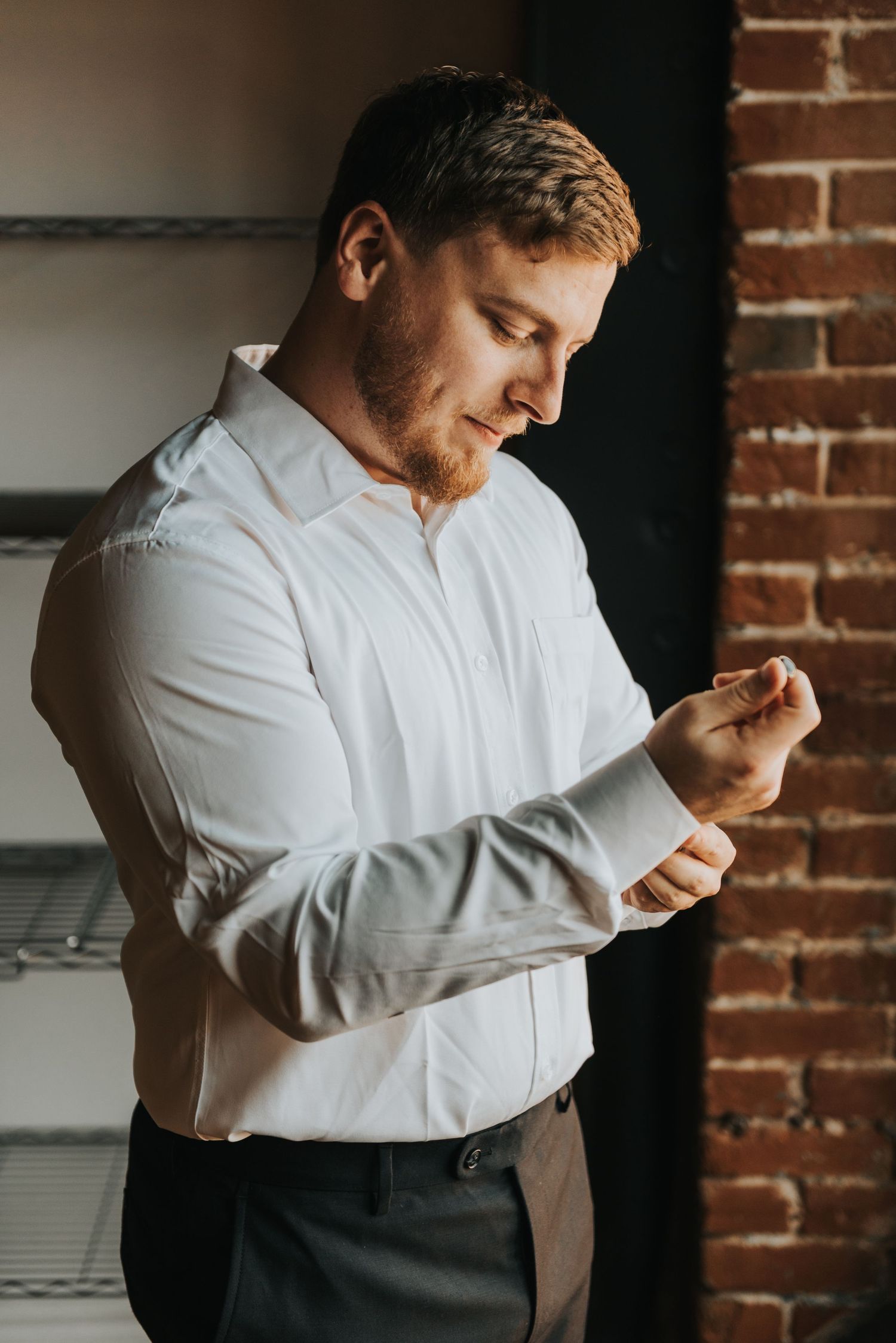 Man in white dress shirt adjusts sleeve cuffs against brick wall background in formal preparation sequence.