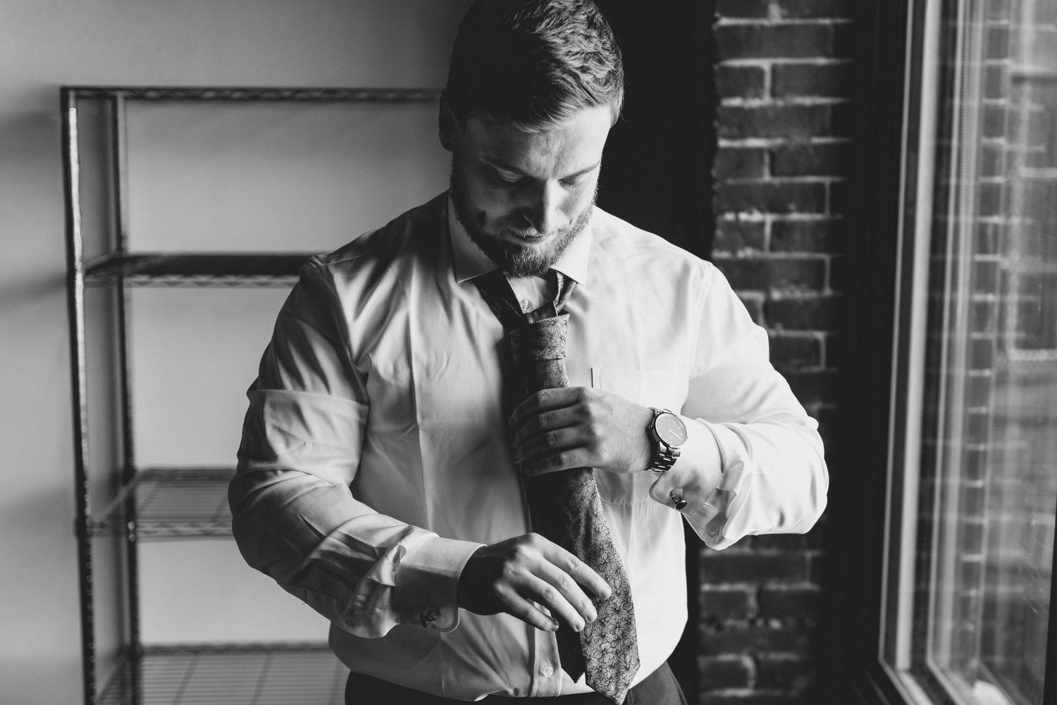 Groomsmen help each other get dressed in formal attire near a brick wall in black and white.