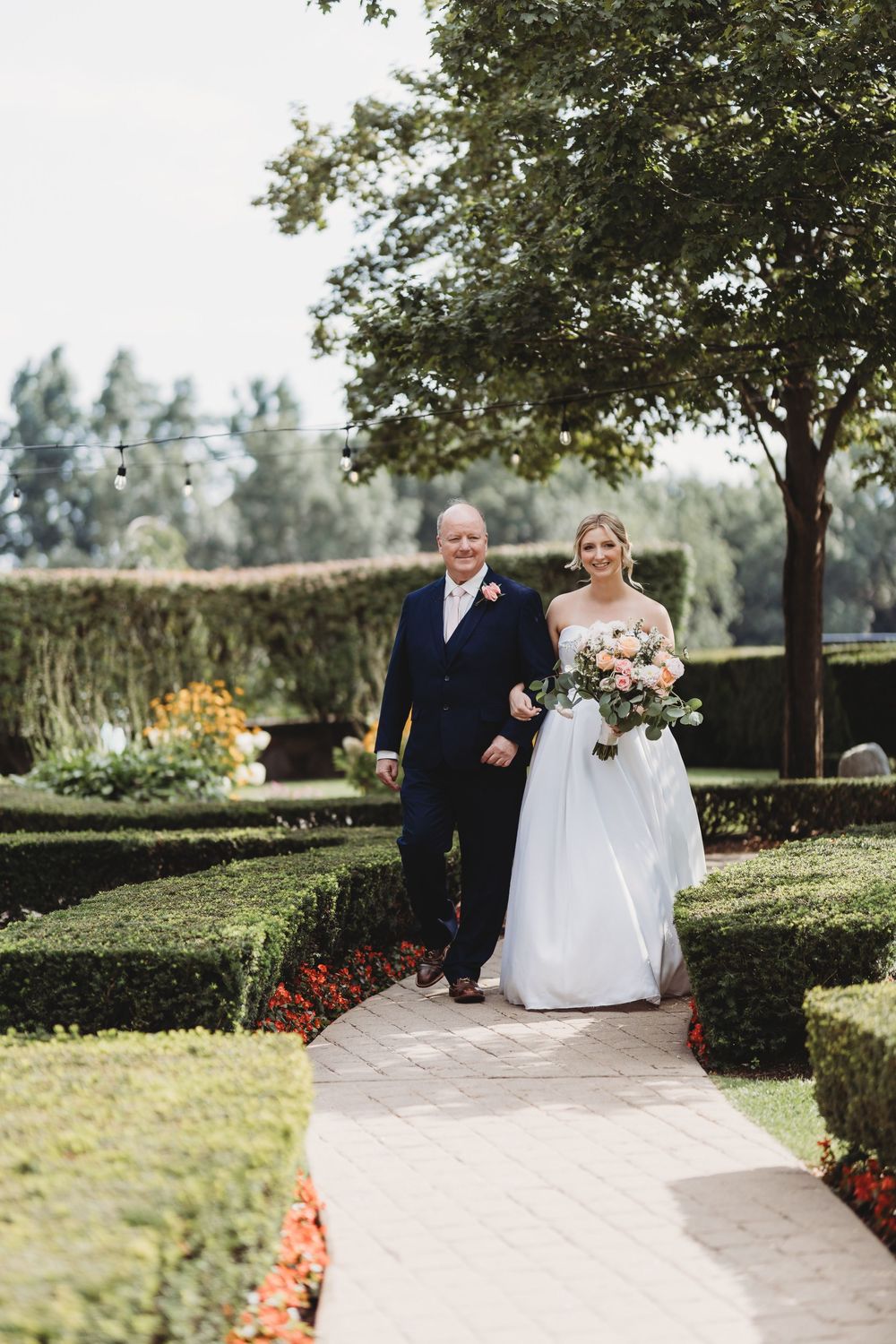 A wedding ceremony in a manicured garden with formal hedges and a stone pathway lined with colorful flowers.