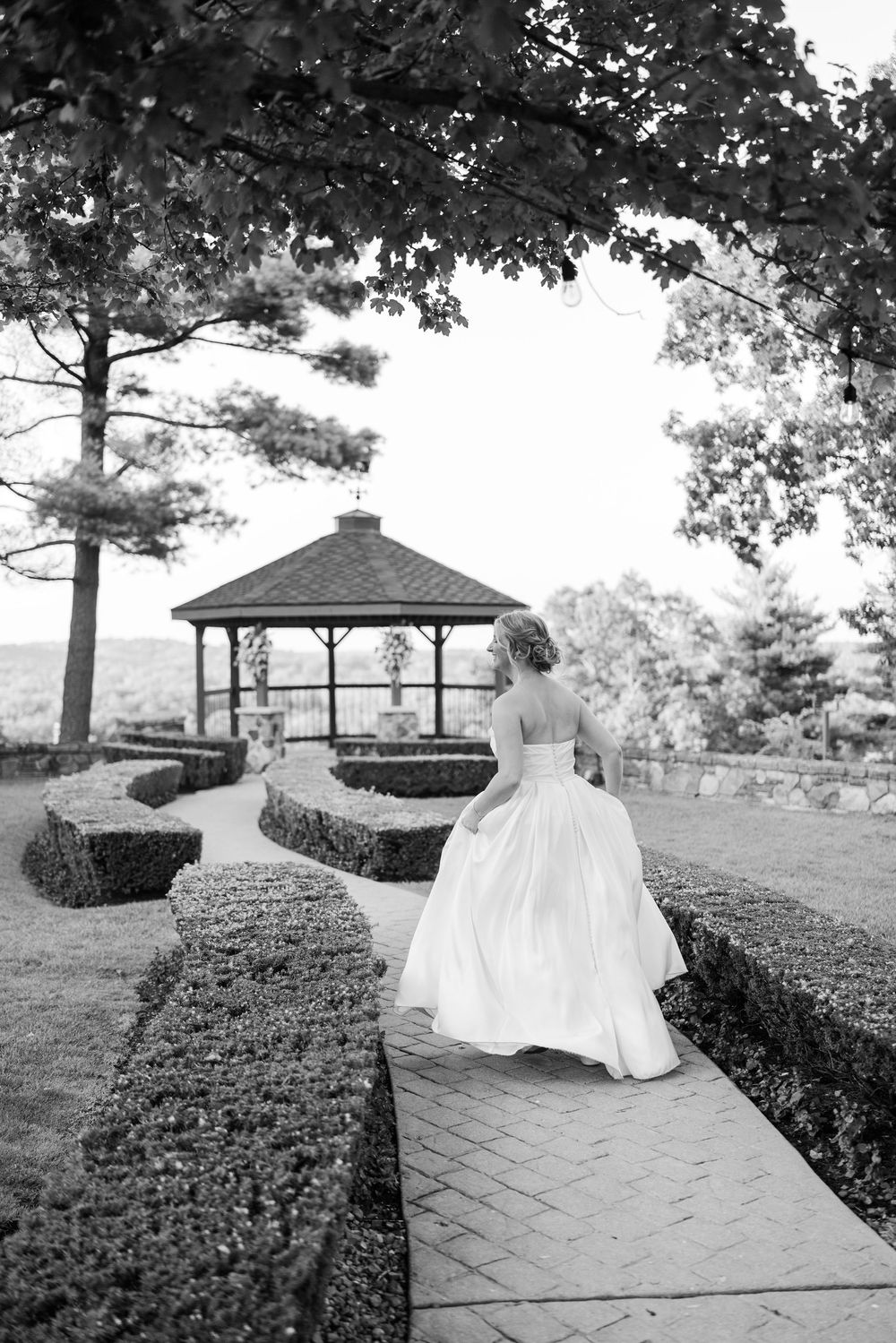 Elegant white wedding dress flowing on a stone path leading to a gazebo in a scenic garden setting.