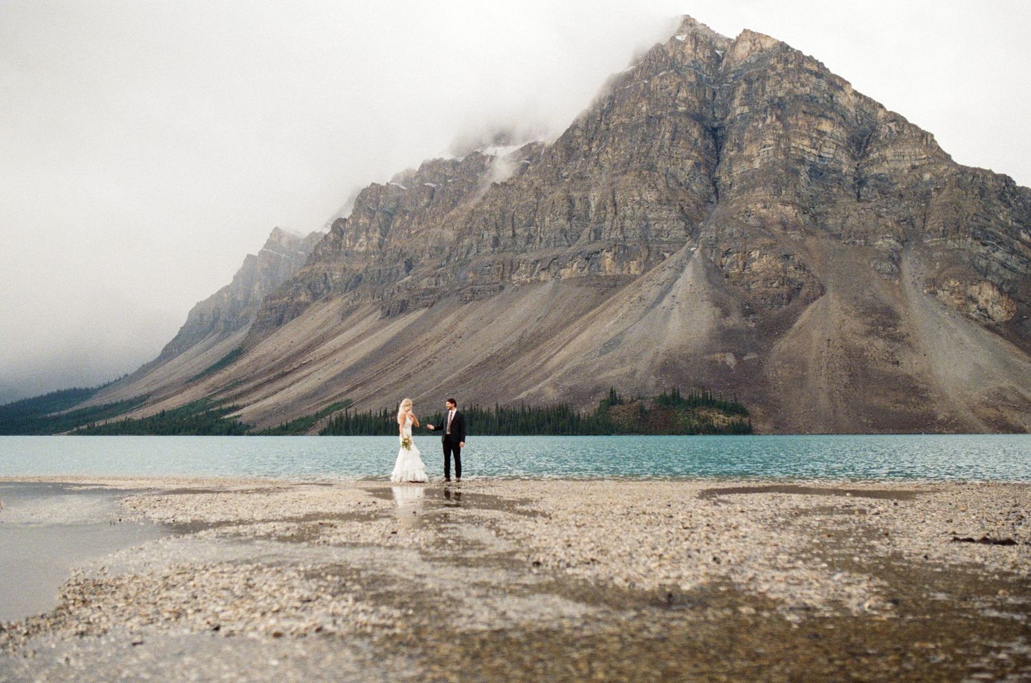 bride and groom walk along the shore of Bow Lake in Banff National Park holding hands