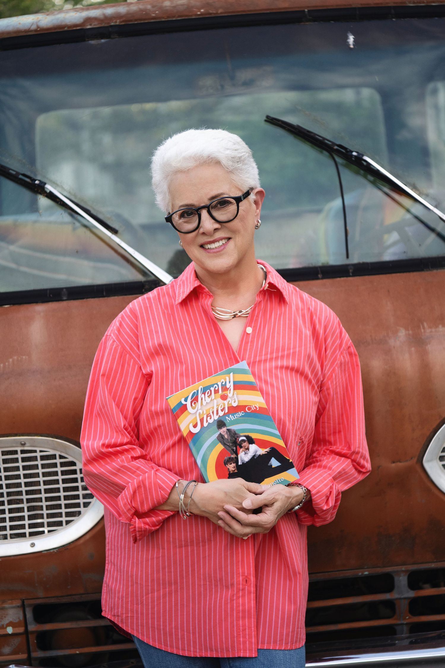 A person in a coral striped shirt holds a colorful rainbow-themed plush toy against a rustic brown car exterior.