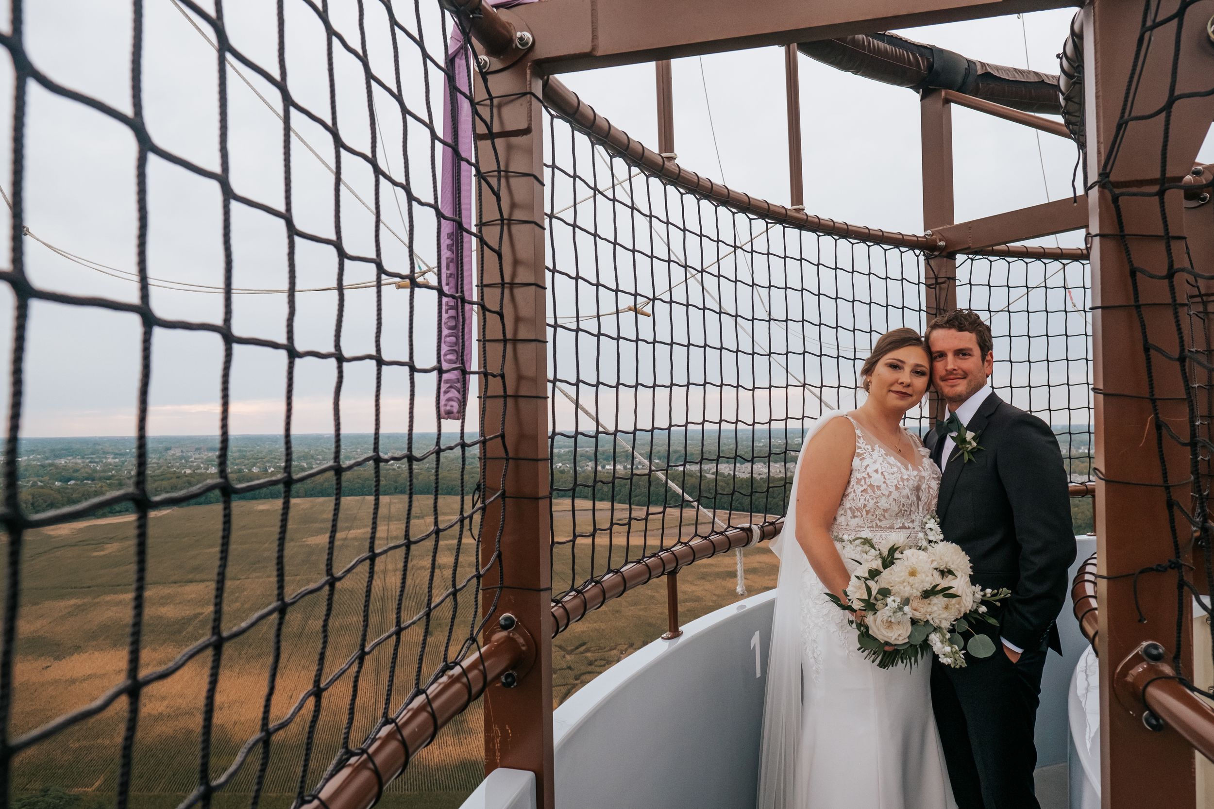 A wedding couple poses in an elevated structure with mesh barriers and scenic views.