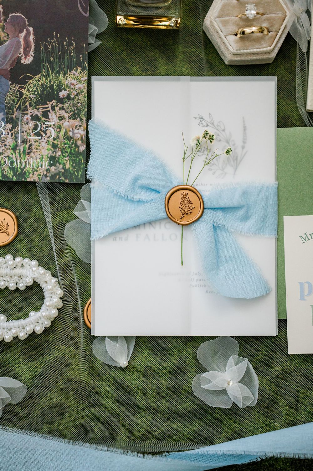 Elegant wedding invitations with light blue ribbons and wax seals displayed on green textured background.