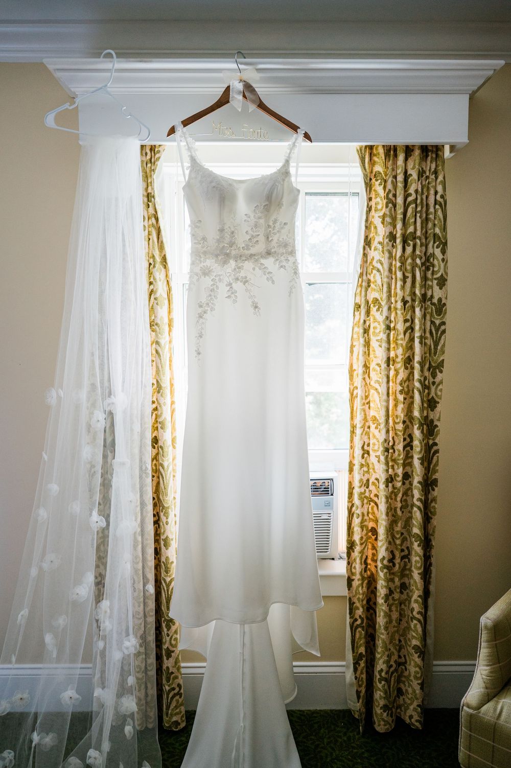 White wedding dress hanging by window with golden floral curtains in natural light.