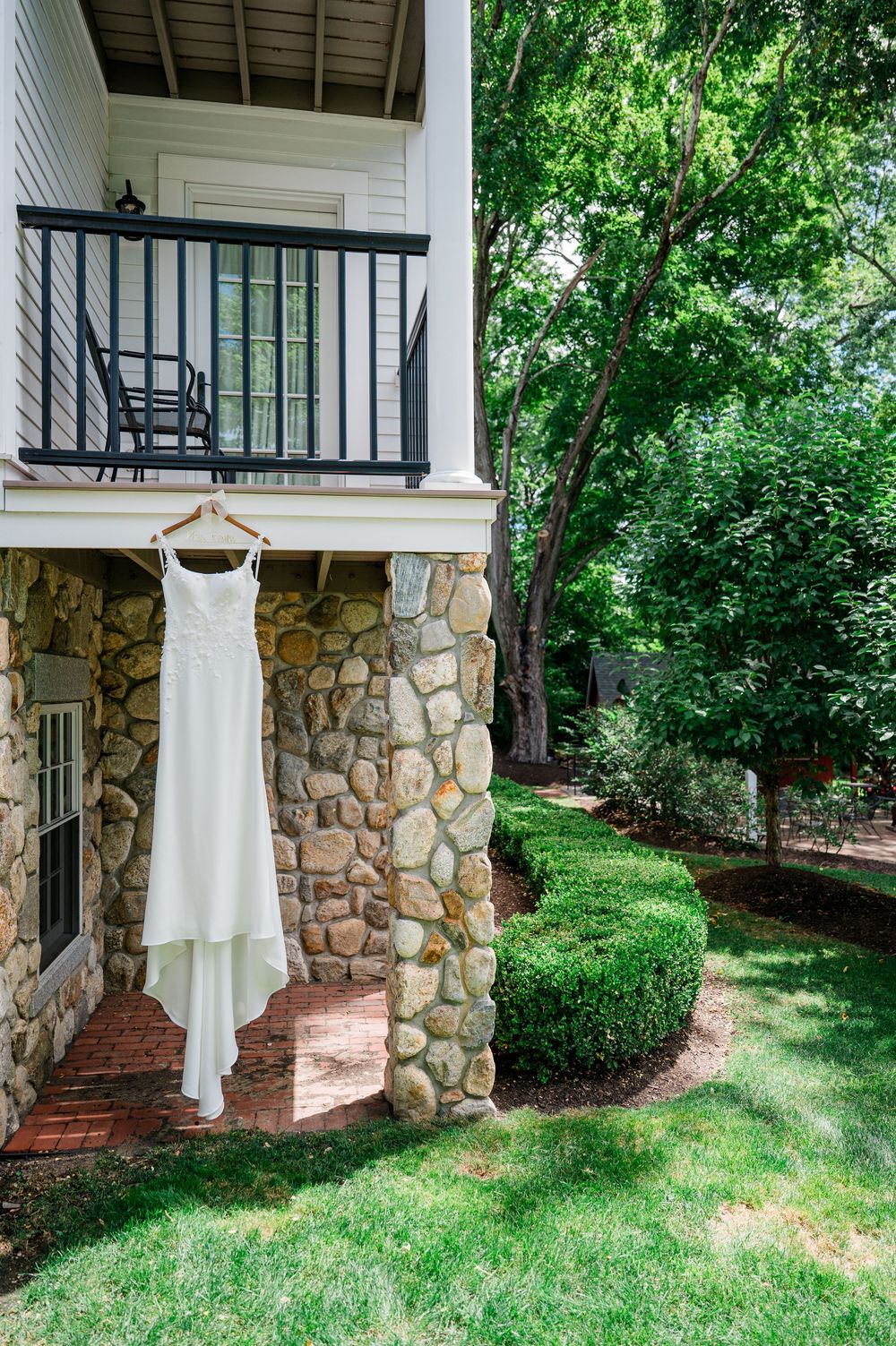 Wedding dress hanging on stone porch with white columns and lush green landscaping.