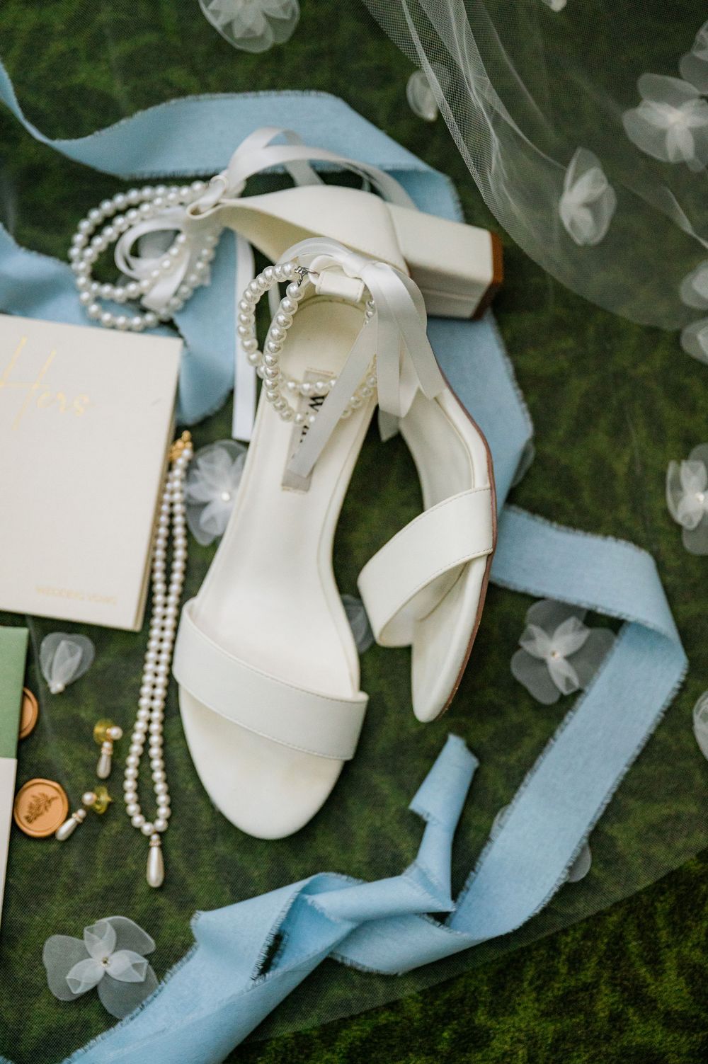 Wedding accessories including white shoes, blue ribbon, and pearl jewelry arranged on grass.