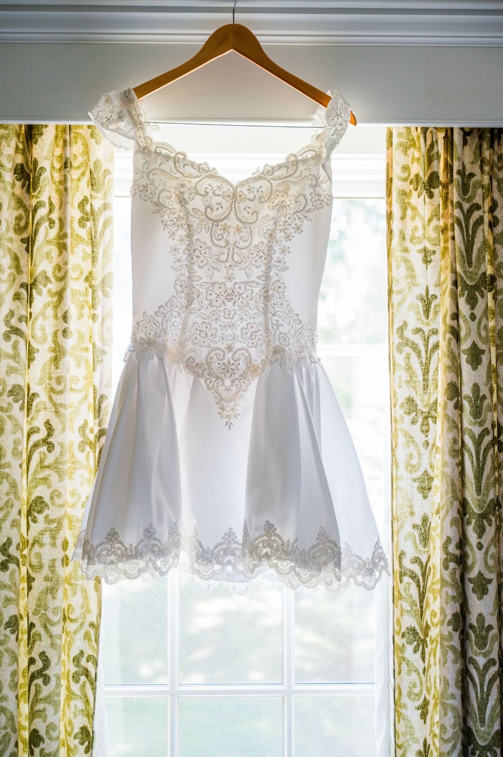 Delicate white lace wedding dresses hanging on hangers against floral curtains in natural light.