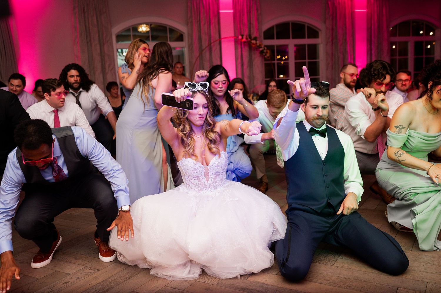 Wedding reception guests dancing energetically on a dance floor illuminated by pink lighting.