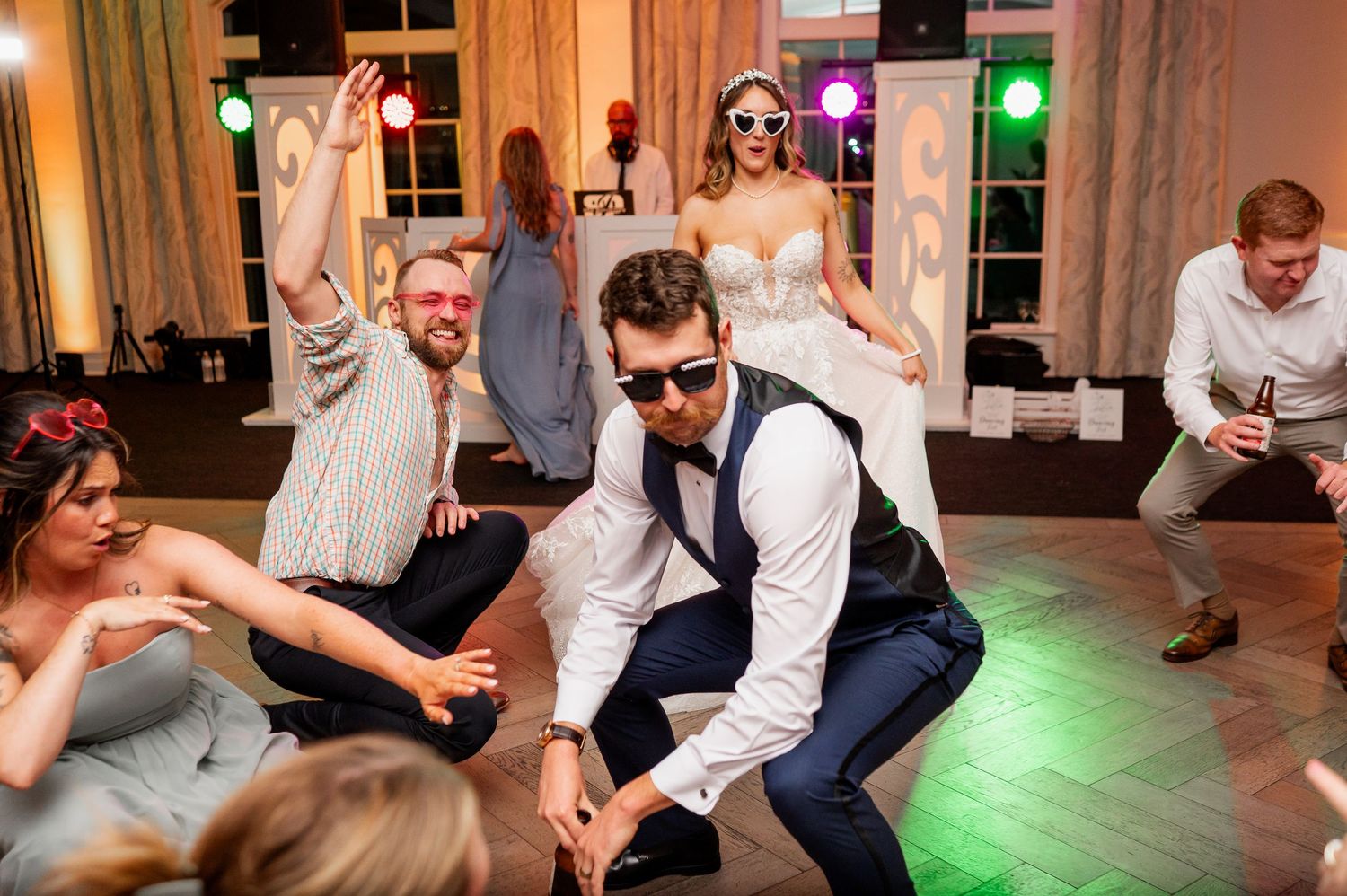 Wedding guests dancing and celebrating in a venue with warm lighting and elegant windows.