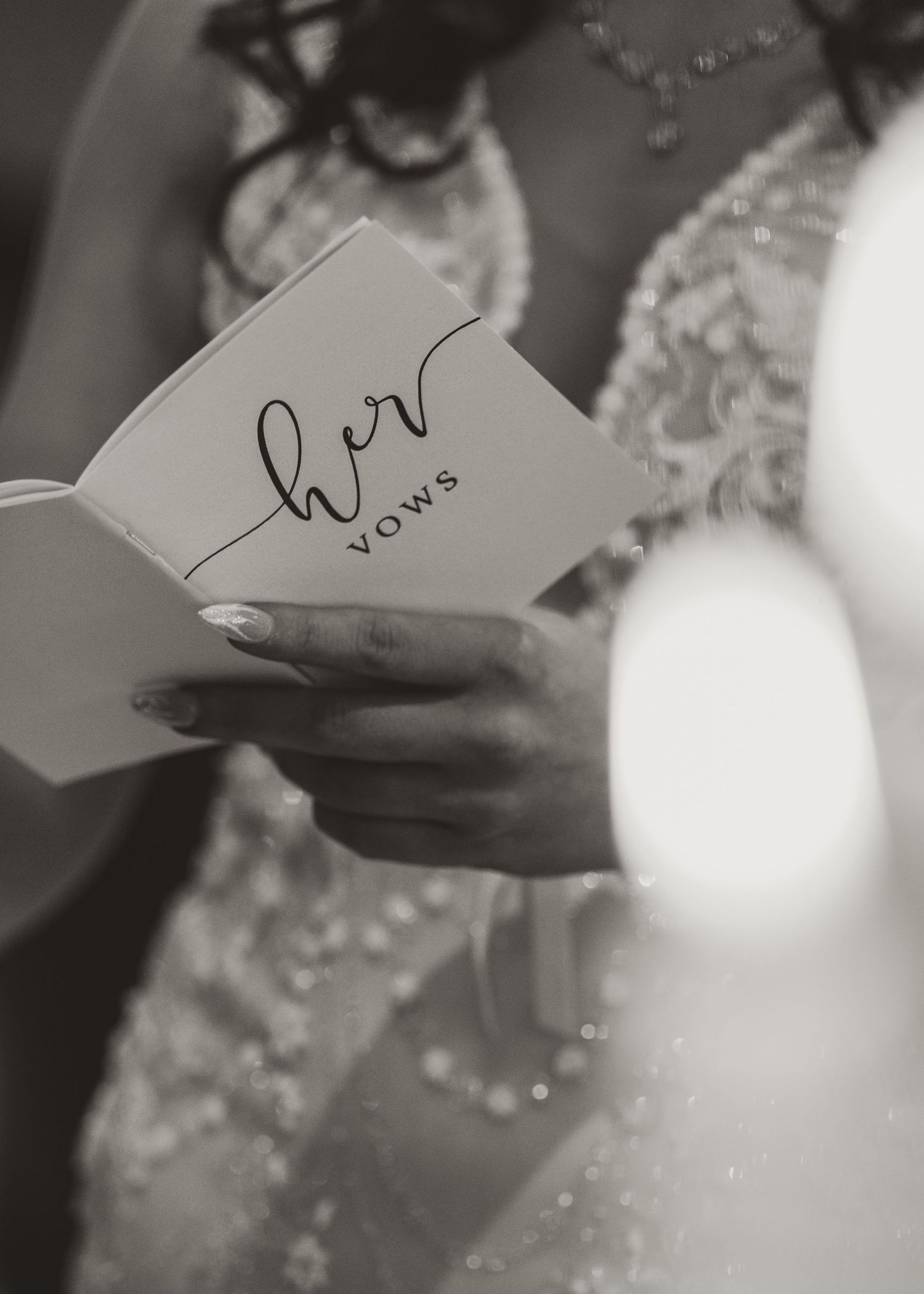 A close-up black and white photo shows hands holding wedding vows written on a white card with elegant script Whitley hall