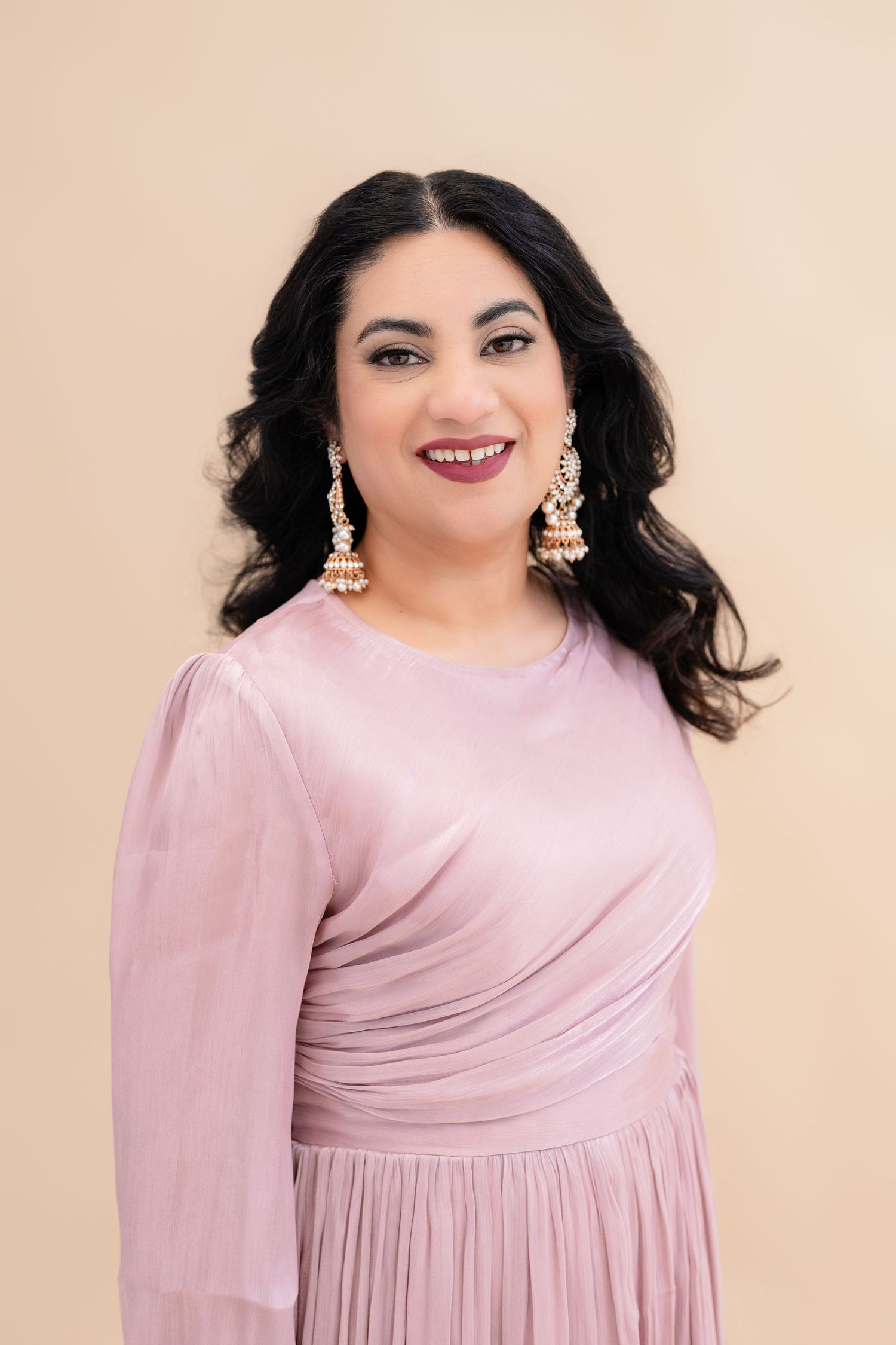 Professional headshot  wearing a soft pink long-sleeved dress with a pleated skirt and statement earrings poses against a beige background.