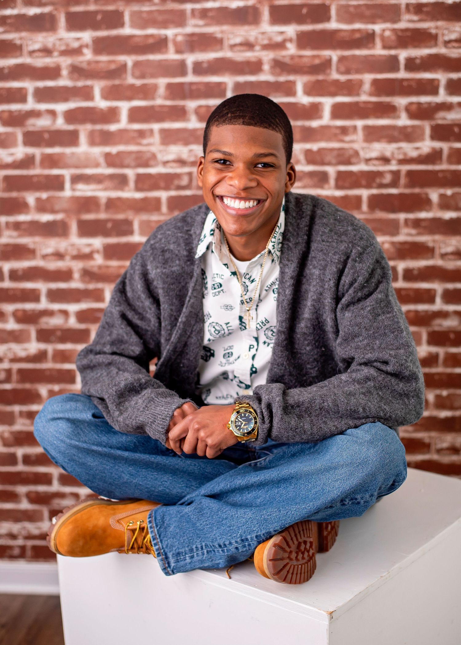 A person in a gray sweater and jeans sits cross-legged against a brick wall, showing a warm smile.