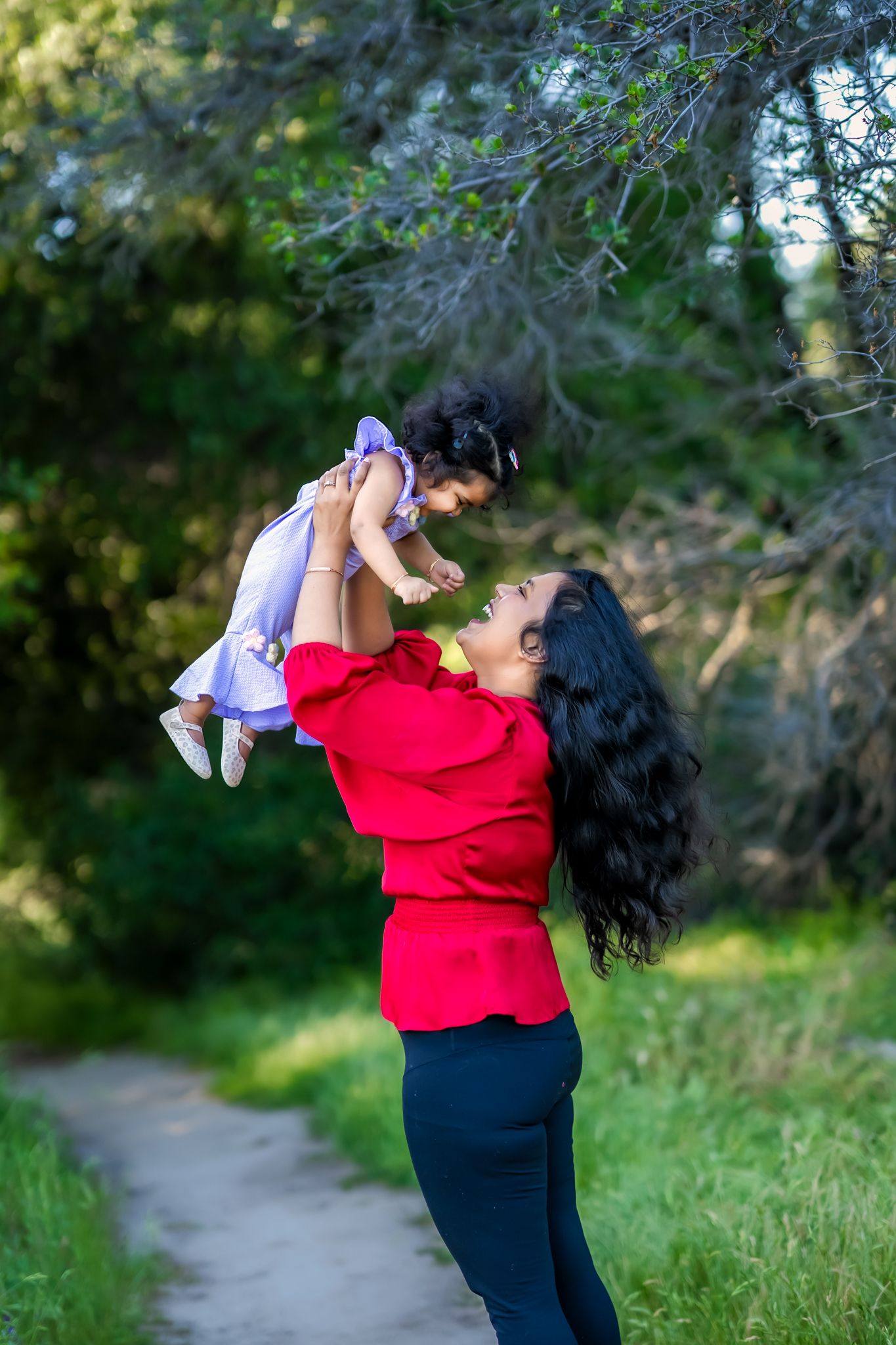 A joyful moment captured on a nature path as a woman in red lifts a small child wearing purple against a green backdrop.