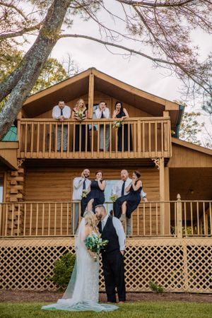 Wedding group strikes fun poses on rustic cabin's wooden balcony.