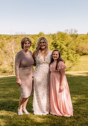Three people wearing pink and beige dresses stand together on a grassy hill during a sunny day.