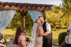 Wedding ceremony gathering under a rustic wooden pergola with flowing blue curtains.